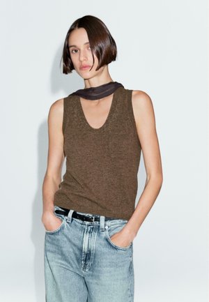 Haut en tricot marron sans manches avec un large décolleté rond, associé à un jean bleu clair taille haute et une ceinture noire. Texture lisse.