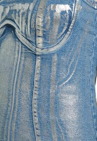 Gros plan sur un tissu en denim bleu avec des coutures piquées et un détail de poche courbée sur un vêtement.