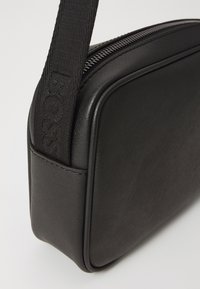 Black rectangular bag made of textured material, featuring a zip closure and a fabric strap embossed with the brand name.