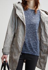 Veste légère grise, résistante à l'eau, avec une capuche, en tissu texturé, dotée de poches avant et d'une taille à cordon de serrage, portée sur un t-shirt en tricot marbré bleu.
