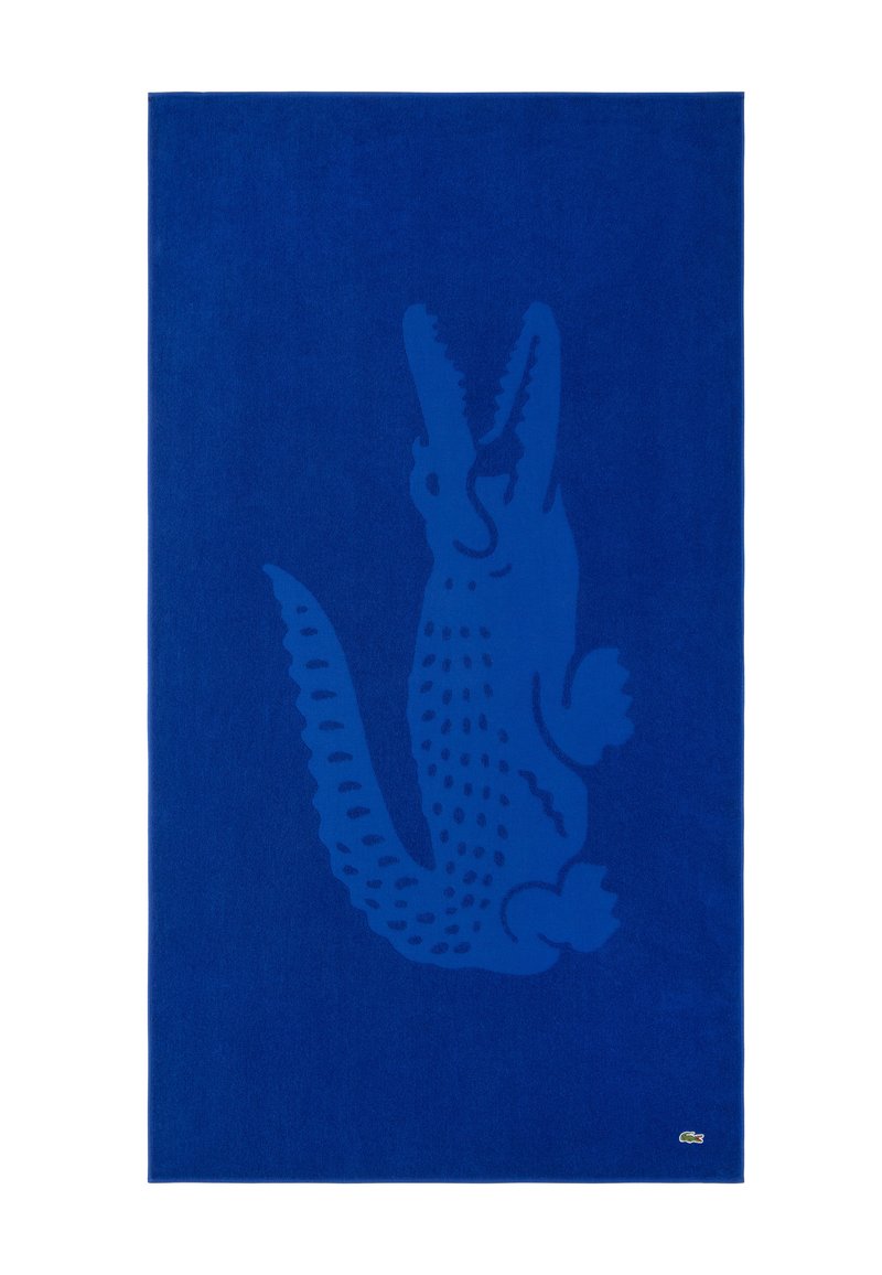 Toalla de baño de algodón azul con un patrón de silueta de cocodrilo en azul más oscuro. Presenta el logo de Lacoste en la esquina inferior. Textura suave.