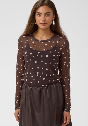 Woman wearing a sheer long-sleeve brown top with beige polka dots over a dark tank top and a dark gathered skirt.