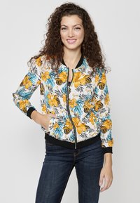 Giacca bomber floreale con base bianca, caratterizzata da fiori gialli e turchesi su fondi neri. Chiusura con zip e tasche laterali.