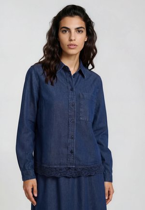 Woman with dark wavy hair wearing a buttoned dark blue denim shirt with a chest pocket and lace trim at the hem, standing against a plain background.