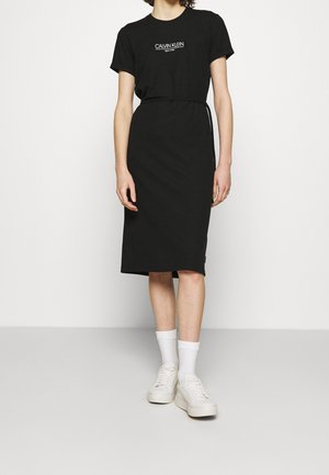 Person iført en sort Calvin Klein t-shirt kjole, hvide sokker og hvide sneakers, stående mod en ensfarvet lys baggrund.