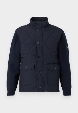 Chaqueta acolchada de color navy con cuello alto, cierre frontal con botones, dos grandes bolsillos y mangas de punto, que presenta una insignia de logo en el brazo izquierdo.