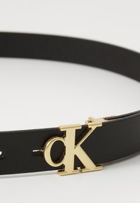 Cintura in pelle nera con un logo "CK" dorato come fibbia. La cintura presenta una texture liscia e un design minimalista.