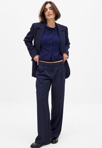 Navy blue tailored blazer over a matching cardigan and high-waisted trousers, featuring a relaxed fit and smooth texture. Black shoes complete the look.