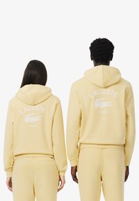 La sudadera amarilla presenta un gran logotipo bordado en blanco de "LACOSTE" y un diseño de cocodrilo en la parte posterior, con capucha ajustable y puños de canalé.