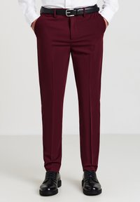 Uomo che indossa pantaloni eleganti bordeaux, cintura nera, scarpe eleganti nere e una camicia bianca con le mani in tasca.