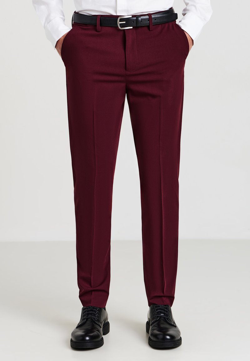 Uomo che indossa pantaloni eleganti bordeaux, cintura nera, scarpe eleganti nere e una camicia bianca con le mani in tasca.