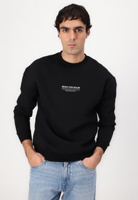 Schwarzer Sweatshirt mit gerippten Bündchen und Ausschnitt, ausgestattet mit einem zentrierten weißen Logo. Kombiniert mit hellblauen Jeans, zeigt einen lässigen Look.