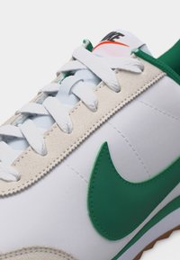 Λευκό αθλητικό παπούτσι με πράσινο σήμα Nike, περιοχή με κορδόνια από μπεζ σουέντ, λευκά κορδόνια και ορατό λογότυπο Nike στη γλώσσα.