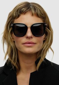 Sorte oversized solbriller med en geometrisk form og mørkt tonede linser. Har guld accenter ved tindingerne, båret med en sort blazer.