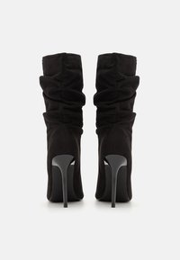 Botas negras de tacón alto hechas de tejido suave, con un diseño fruncido en la caña y un tacón elegante y brillante.