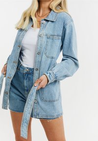 Chaqueta de mezclilla azul claro con cierre de botones, dos bolsillos en el pecho y cintura ceñida. Mangas largas con deshilachado sutil en el dobladillo.