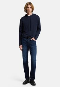 Felpa navy con coulisse, abbinata a jeans blu scuri. Modello che indossa scarpe nere lucide, in piedi su uno sfondo neutro.