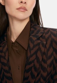 Brown blazer with a zigzag pattern in darker brown, featuring a notched collar and a brown shirt underneath, secured with a button.