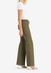 Jean large kaki avec poches avant, associé à un haut blanc à manches courtes côtelé et des sandales blanches à talons, vu de profil.