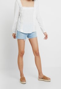 Blouse blanche à manches longues avec décolleté carré et bordure en dentelle, associée à un short en denim bleu clair et des sandales brunes à enfiler avec une semelle en jute.