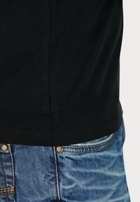 Čierne bavlnené tričko s okrúhlym výstrihom, skombinované s modrými džínsami z denimu s vyblednutým dizajnom a kontrastným prešívaním. Viditeľný detail s kovovým gombíkom.