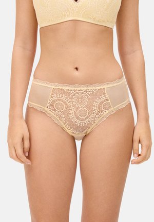 Braguitas de encaje beige de talle alto con patrones florales y paneles laterales de malla, llevadas por una persona de pie contra un fondo claro y liso.