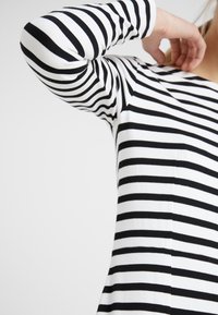 Osoba w czarno-białą pasiastą koszulę z długim rękawem, z uniesioną ręką blisko twarzy na jednolitym tle.