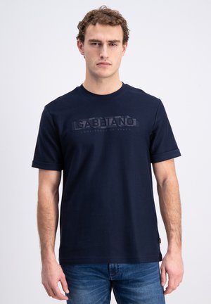 Młody mężczyzna z kręconymi włosami nosi ciemnoniebieski t-shirt Gabbiano i niebieskie dżinsy, stoi na jasnym, jednolitym tle.