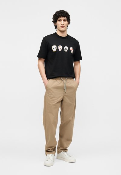 Νεαρός άνδρας στέκεται με τα χέρια στις τσέπες, φοράει μαύρο μπλουζάκι με τέσσερα πολύχρωμα σχέδια κρανίων, μπεζ παντελόνι και λευκά αθλητικά παπούτσια.