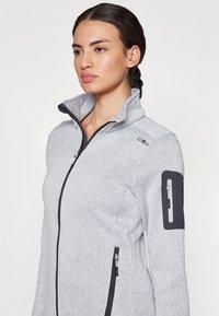 Veste en polaire grise à fermeture éclair avec col haut, dotée d'une poche latérale et d'accents noirs. Texture douce avec un motif subtil sur l'ensemble.