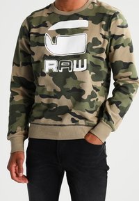Felpa camouflage in toni di verde e beige, con un logo e una grafica "RAW" bianchi. Maniche e orlo a costine offrono texture e vestibilità.
