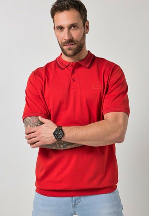 Mann mit Bart und Tätowierungen trägt ein rotes Poloshirt und eine schwarze Armbanduhr, steht mit verschränkten Armen vor einfachem Hintergrund.
