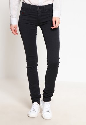 Osoba nosząca obcisłe czarne dżinsy, białe trampki i białą koszulę z długim rękawem, stojąca na jasnym tle.