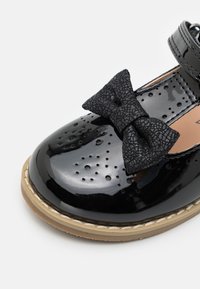 Zapato de charol negro con diseño perforado, puntera redonda y un detalle de lazo negro texturizado. Suela teñida con un ligero detalle de costura.