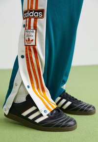 Černé boty Adidas s třemi bílými pruhy a červenými detaily. Tyrkysové tepláky s bílými a oranžovými bočními panely a zapínáním na druky.