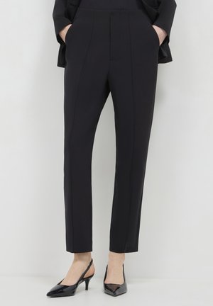 Woman wearing tailored black ankle-length pants with seam details, hands in pockets, paired with black pointed-toe slingback heels.