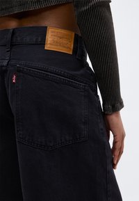 Widok z tyłu osoby noszącej czarne jeansy Levi's oraz prążkowany, ciemnozielony top z długim rękawem, z widoczną skórzaną naszywką Levi's i czerwonym tagiem.