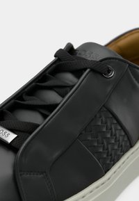 Zbliżenie na czarny skórzany sneaker z plecionym bocznym panelem, czarnymi sznurówkami i białą podeszwą, ukazujące szczegółowe przeszycia i metalową plakietkę z logo.