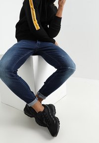 Jeans in denim blu con una vestibilità affusolata e leggeri segni di usura sulle ginocchia, abbinati a una felpa nera con dettagli gialli e sneakers nere.