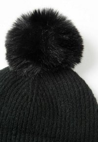 Melna adīta cepure ar ribotu tekstūru, kurai ir liels melns mākslīgā kažoka pom-poms augšpusē. Tuvplāns uzsver detaļas.