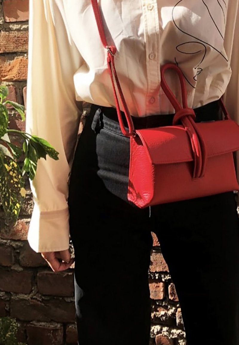 Sac à bandoulière en cuir rouge avec une texture lisse, un design minimaliste et un accent noué. Porté avec une chemise blanche et un pantalon noir.