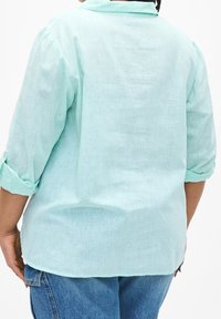 Lehká světle zelená lněná košile se stojáčkem, vyhrnutými rukávy a volným střihem, s texturovaným povrchem. Na zadní straně je rovný lem.