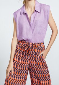 Lila ärmlös blus med krage och ficka, tillsammans med vida byxor med ett orange och marinblått vågmönster, åtdragna vid midjan.