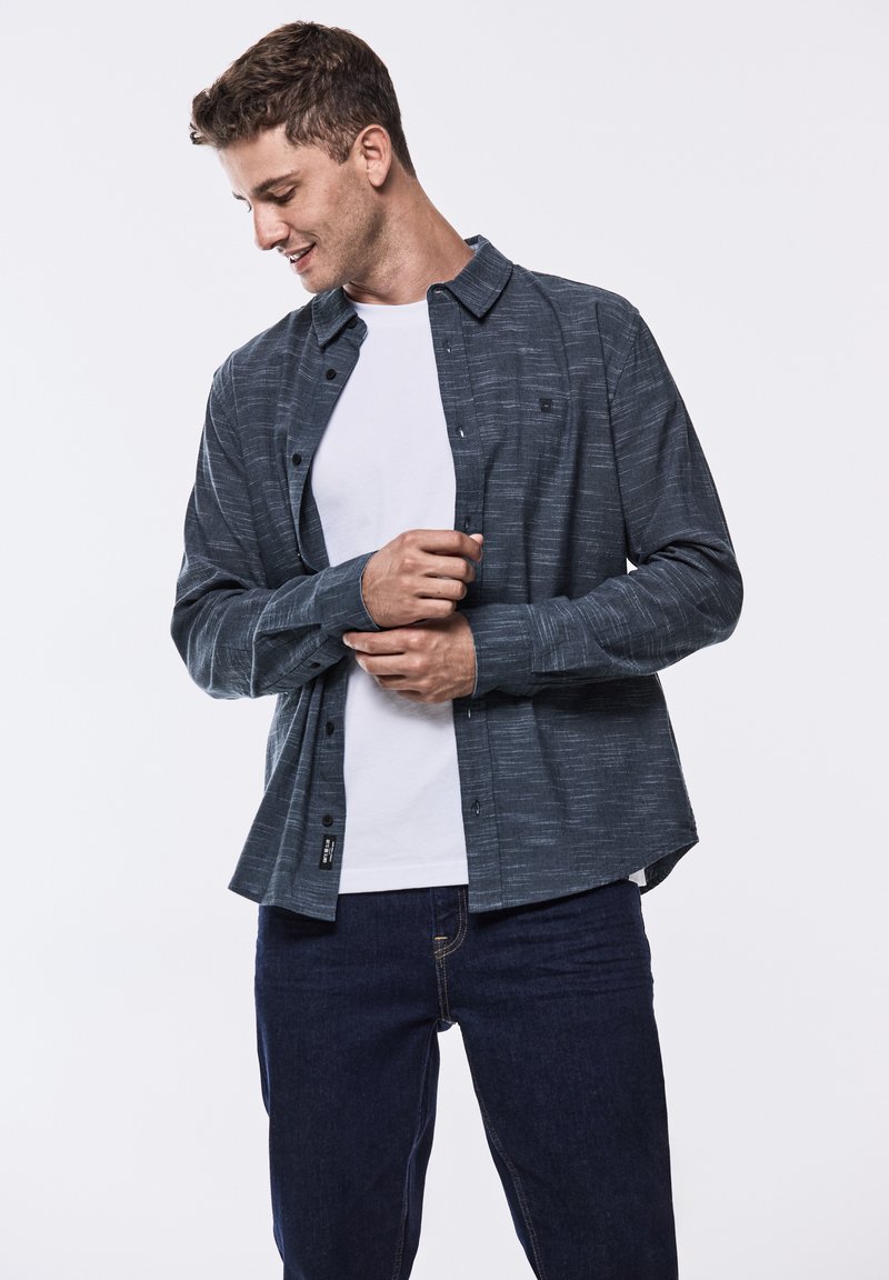 Camicia button-up blu scuro con un motivo a trama, indossata sopra una t-shirt bianca, abbinata a jeans scuri. Presenta una vestibilità casual e maniche arrotolate.