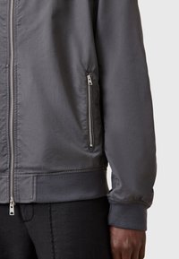Giacca bomber grigia con una texture liscia e opaca, chiusura frontale con zip e tasche laterali con hardware argentato; polsini e orlo a costine.