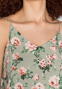Débardeur floral vert avec des roses rose clair et blanches, décolleté en V et fines bretelles, fabriqué à partir d'un tissu doux au toucher lisse.