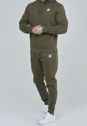 Mann trägt olivgrünen Hoodie und passende Jogginghose mit weißem Logo, steht mit verschränkten Händen, weiße Turnschuhe vor hellem Hintergrund.