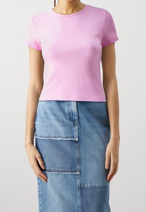 Femme portant un haut rose ajusté à manches courtes et une jupe en jean patchwork aux bords effilochés, debout devant un fond uni.