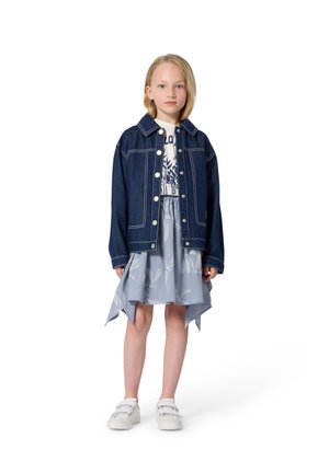 Spijkerjack met witte stiksels, knoopsluiting, over een grafisch T-shirt en een lichtblauwe rok met bloemenpatroon; witte sneakers.