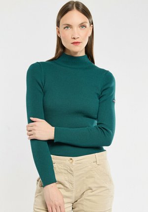 Teal geribbelde coltrui met lange mouwen. Zachte textuur en aansluitend design. G gedragen met lichtbeige broek. Opvallend logo op de mouw.